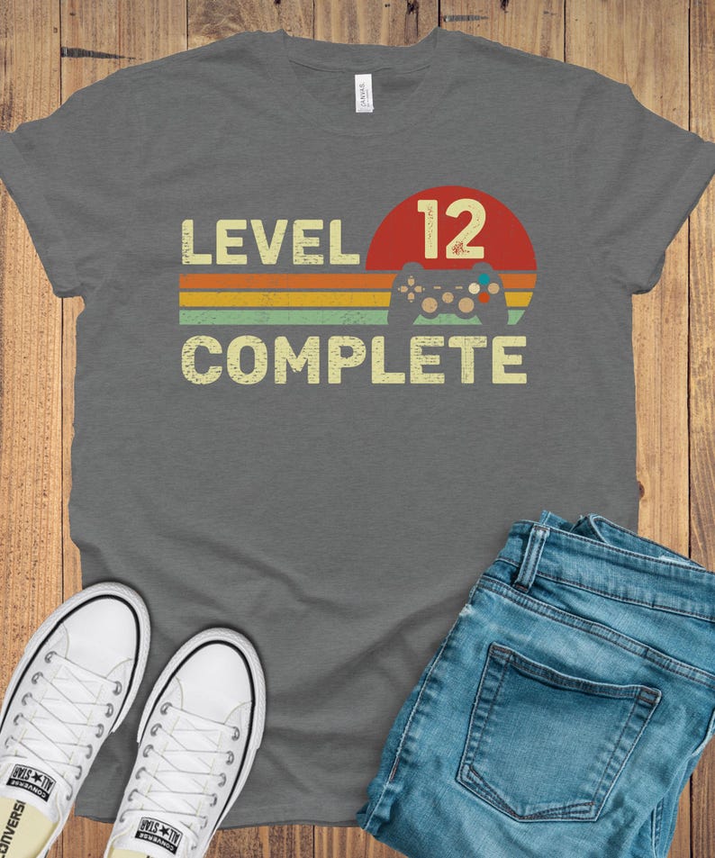 May include: A gray t-shirt with the text "LEVEL 12 COMPLETE" in a retro font. The design includes a red circle with the number 12, a controller graphic, and colorful stripes. White sneakers and blue jeans are also visible.