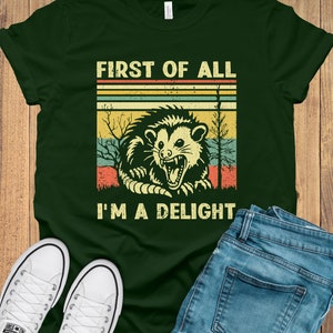 May include: A green t-shirt with a vintage-style graphic of a possum with the text "First of all I'm a delight".