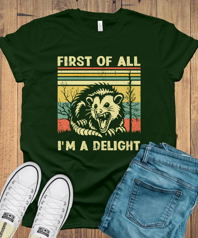 May include: A green t-shirt with a vintage-style graphic of a possum with the text "First of all I'm a delight".