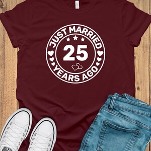 May include: Burgundy t-shirt with a white circular graphic that reads "JUST MARRIED 25 YEARS AGO." The shirt is paired with blue jeans and white canvas sneakers. The design includes stars and hearts.