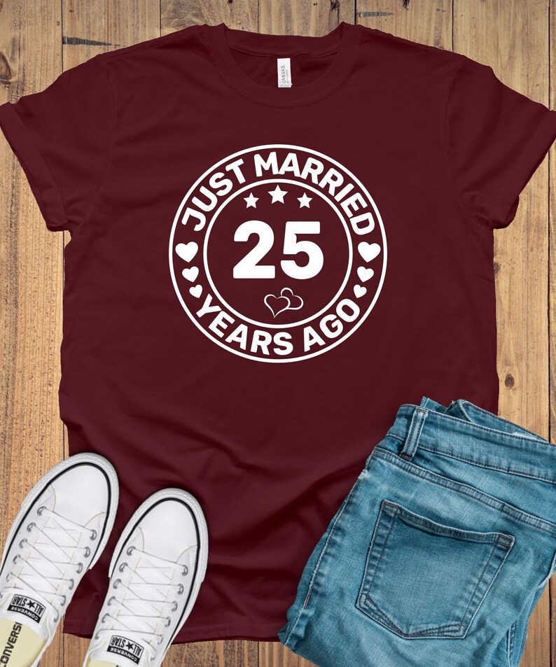 May include: Burgundy t-shirt with a white circular graphic that reads "JUST MARRIED 25 YEARS AGO." The shirt is paired with blue jeans and white canvas sneakers. The design includes stars and hearts.