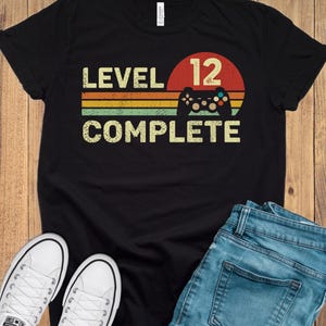 May include: Black t-shirt with the text "LEVEL 12 COMPLETE" in a retro design. The design includes a controller graphic and a sunset-style background with orange, yellow, and green stripes. White sneakers and blue jeans are also visible.