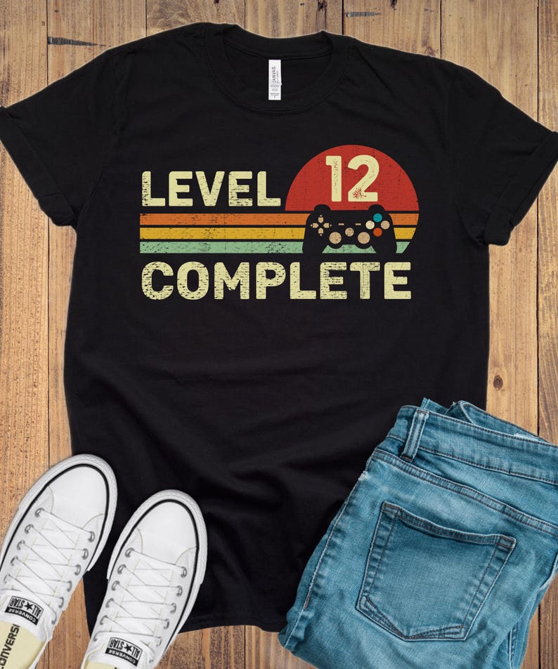 May include: Black t-shirt with the text "LEVEL 12 COMPLETE" in a retro design. The design includes a controller graphic and a sunset-style background with orange, yellow, and green stripes. White sneakers and blue jeans are also visible.