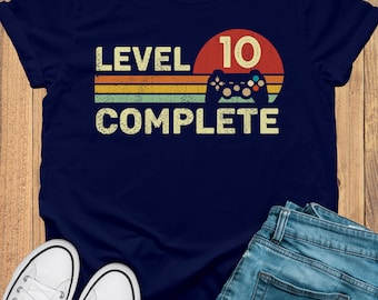 May include: Navy blue t-shirt with the text "LEVEL 10 COMPLETE" in a retro design. The design includes a controller graphic and a red circle. The shirt is paired with blue jeans and white sneakers.