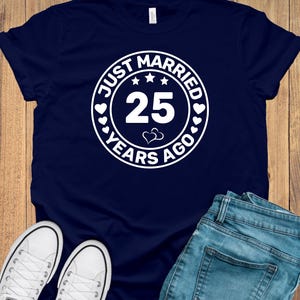 May include: Navy blue t-shirt with a white circular graphic that reads "JUST MARRIED 25 YEARS AGO" with stars and hearts. The image also shows white sneakers and blue jeans.