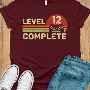 May include: A maroon t-shirt with the words "LEVEL 12 COMPLETE" in a retro font, with a controller graphic. The design is set against a red and orange sunset-style background. White sneakers and blue jeans are also in the image.