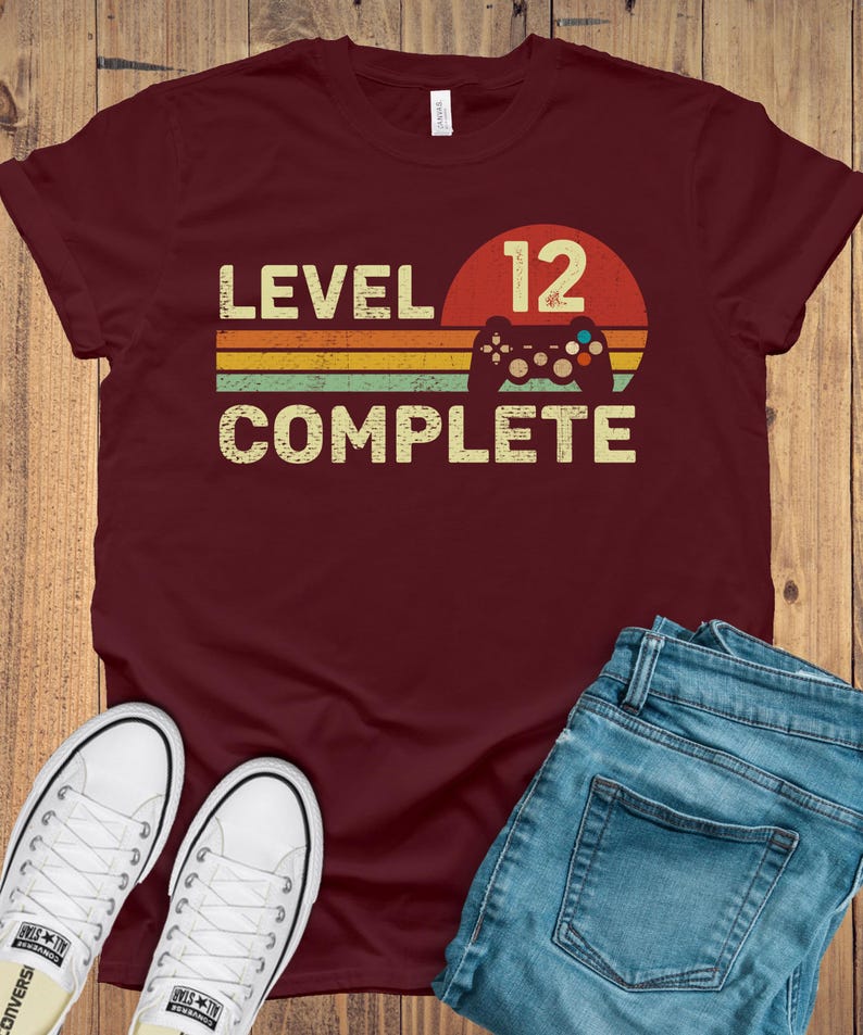 May include: A maroon t-shirt with the words "LEVEL 12 COMPLETE" in a retro font, with a controller graphic. The design is set against a red and orange sunset-style background. White sneakers and blue jeans are also in the image.