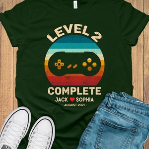 Puede incluir: Una camiseta verde con un gr&aacute;fico de mando de videojuego retro y el texto "Level 2 Complete Jack &hearts; Sophia - August 2021 - "