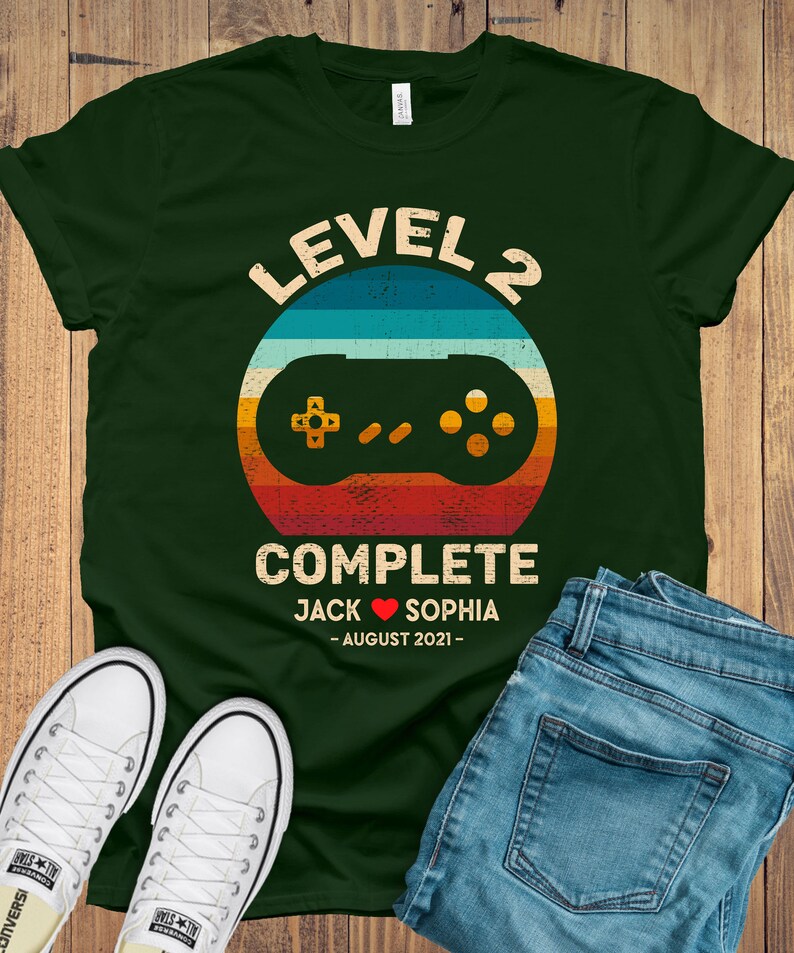 Puede incluir: Una camiseta verde con un gr&aacute;fico de mando de videojuego retro y el texto "Level 2 Complete Jack &hearts; Sophia - August 2021 - "