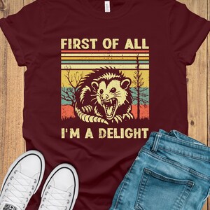 May include: A maroon t-shirt with a vintage-style graphic of a possum with the text "First of all I'm a delight".