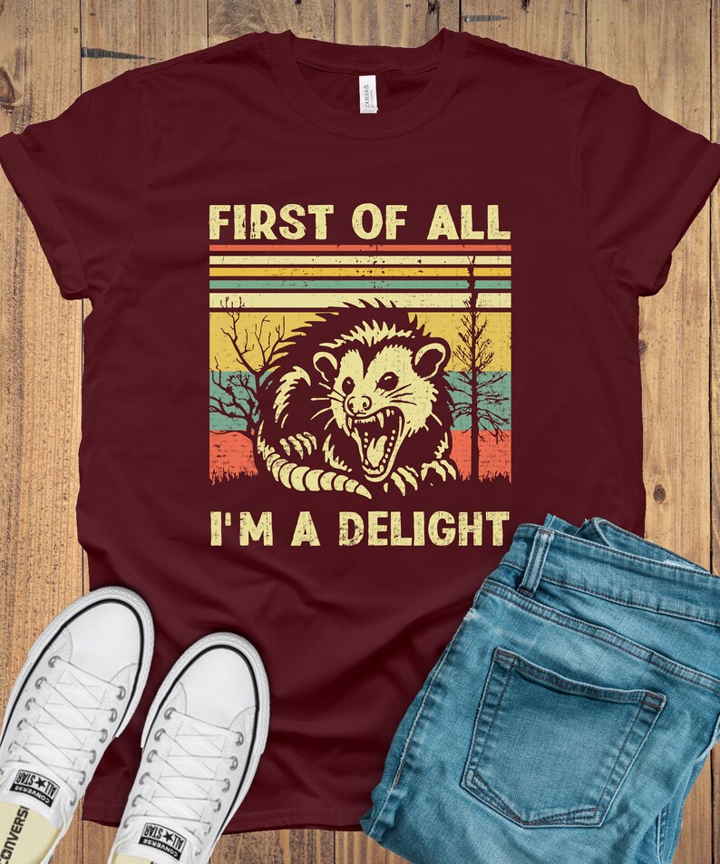 May include: A maroon t-shirt with a vintage-style graphic of a possum with the text "First of all I'm a delight".