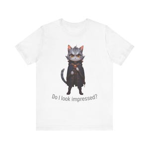 Puede incluir: Camiseta blanca con un gato de dibujos animados vestido con una capa oscura y botas, sosteniendo un orbe dorado. El gato tiene pelaje gris y ojos amarillos. El texto dice: "Do I look impressed?"