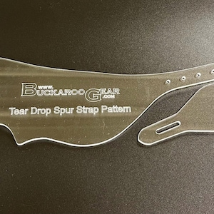Puede incluir: Patrón de correa de espuela Tear Drop de acrílico transparente con el texto "Buckaroo Gear.com". El patrón incluye una pieza larga y curvada con una correa y una pieza más pequeña en forma de lágrima.