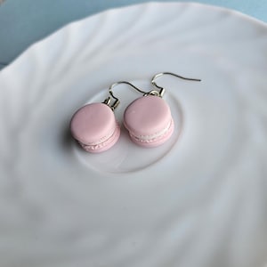 Peut inclure: Deux boucles d'oreilles macarons roses avec des crochets argentés, posées sur une assiette blanche. Les boucles d'oreilles sont rose clair avec un remplissage blanc, ressemblant à de petits desserts. L'assiette blanche présente un motif ondulé.