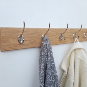 May include: A wooden coat rack with four silver hooks. Two items of clothing are hanging on the hooks, one is a white sweater and the other is a grey patterned scarf.