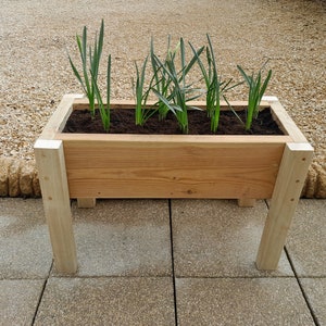 May include: A wooden planter box with four legs, filled with soil and green plants. The planter is sitting on a patio.