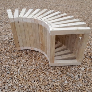 May include: A curved wooden bench constructed from light-coloured timber. The bench features a slatted back and a solid, curved base. The design is unique and the construction appears robust, suitable for outdoor use. The bench is set on a gravel surface.