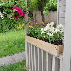 May include: A wooden planter box with white flowers and green plants, attached to a white wooden railing. The planter box is mounted on the side of a white wooden shed.