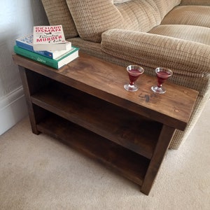 May include: A dark brown wooden side table with two shelves. The table has a distressed finish and is next to a beige sofa. Two glasses of red wine are on the table.