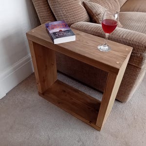 May include: A wooden side table with a book and a glass of red wine on top. The book is titled "Kingdom of the Blind". The table is made of light brown wood and has a hollow centre. The table is next to a beige sofa.