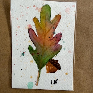 Könnte beinhalten: Ein Aquarellbild auf strukturiertem weißem Papier mit einem einzelnen Eichenblatt, das von Grün zu Orange und Violett übergeht. Eine kleine braune Eichel befindet sich am Blattansatz. Der Hintergrund ist mit bunten Farbspritzern und den Initialen LW verziert.