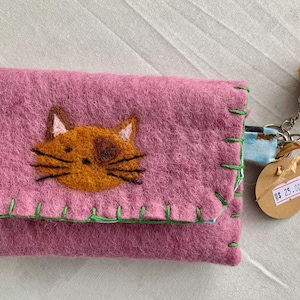 May include: A pink felted wallet featuring a stitched cat face in shades of brown and tan. The wallet has bright green whip-stitching along the edges and includes a brown pom-pom keychain with a circular tag displaying the text $ 25.00.