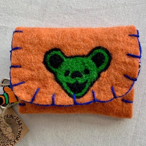 May include: Orange felt wallet with a green bear face embroidered on the front. The wallet has blue stitching around the edges and a silver metal ring attached to a colorful fabric strap.