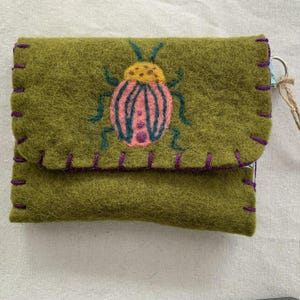 Puede incluir: Cartera de fieltro verde oliva con cierre de solapa, cosida a mano con hilo morado. La parte delantera presenta un diseño de insecto bordado en rosa, amarillo y verde azulado. Incluye un pequeño anillo de metal y una cuerda de fibra natural.