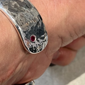 May include: A silver hammered cuff bracelet with a single red gemstone set in the centre.