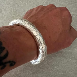 May include: A thick, silver-coloured bangle bracelet featuring a distinct hammered texture across its entire surface. The metal has a reflective, polished finish that catches the light, creating a series of small, irregular indentations.