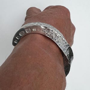 May include: A silver cuff bracelet featuring a hammered, textured surface. The interior of the band displays clear hallmarks, including the number 925, indicating sterling silver composition. The metal has a polished, reflective finish throughout.