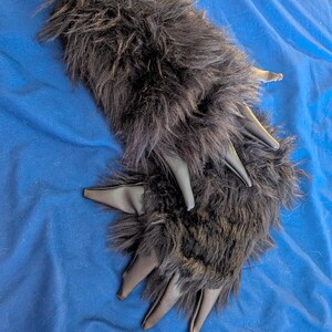 May include: A pair of black faux fur gloves featuring long, pointed, smooth black synthetic leather claws extending from the fingertips. The gloves are covered in thick, shaggy black faux fur that covers the entire back of the hand and fingers.