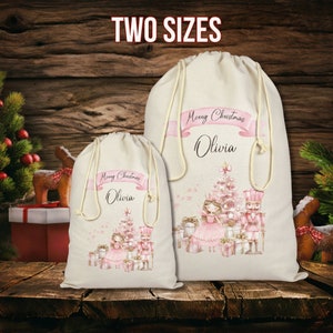 May include: Two beige drawstring Christmas gift bags with a pink and white design. The bags feature a Christmas tree, a ballerina, a nutcracker, and the text "Merry Christmas Olivia". The words "TWO SIZES" are at the top.
