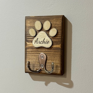 May include: Wooden dog lead hook with a paw print design and the name "Archie" engraved on it. The hook is brown and has a silver metal hook.