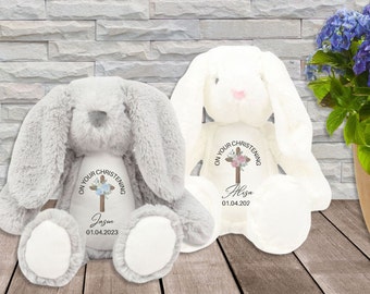 May include: Two plush gray and white bunny rabbits with embroidered crosses and floral designs. The text "On your christening" is embroidered on each bunny. One bunny has the name "Jason" and the date "01.04.2023" embroidered on it. The other bunny has the name "Eliza" and the date "01.04.202" embroidered on it.
