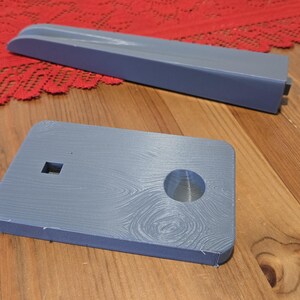 May include: A gray 3D printed phone stand with a circular hole in the center and a small square cutout. The stand is on a wooden surface.