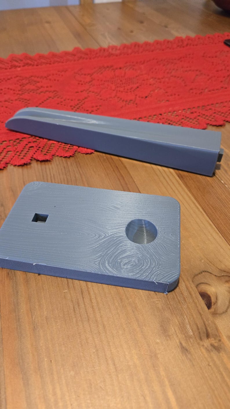 May include: A gray 3D printed phone stand with a circular hole in the center and a small square cutout. The stand is on a wooden surface.