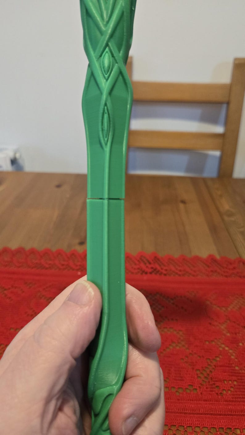 May include: A green plastic staff with a detailed design of intertwined vines and leaves. The staff is split in two pieces, showing the inside of the staff.