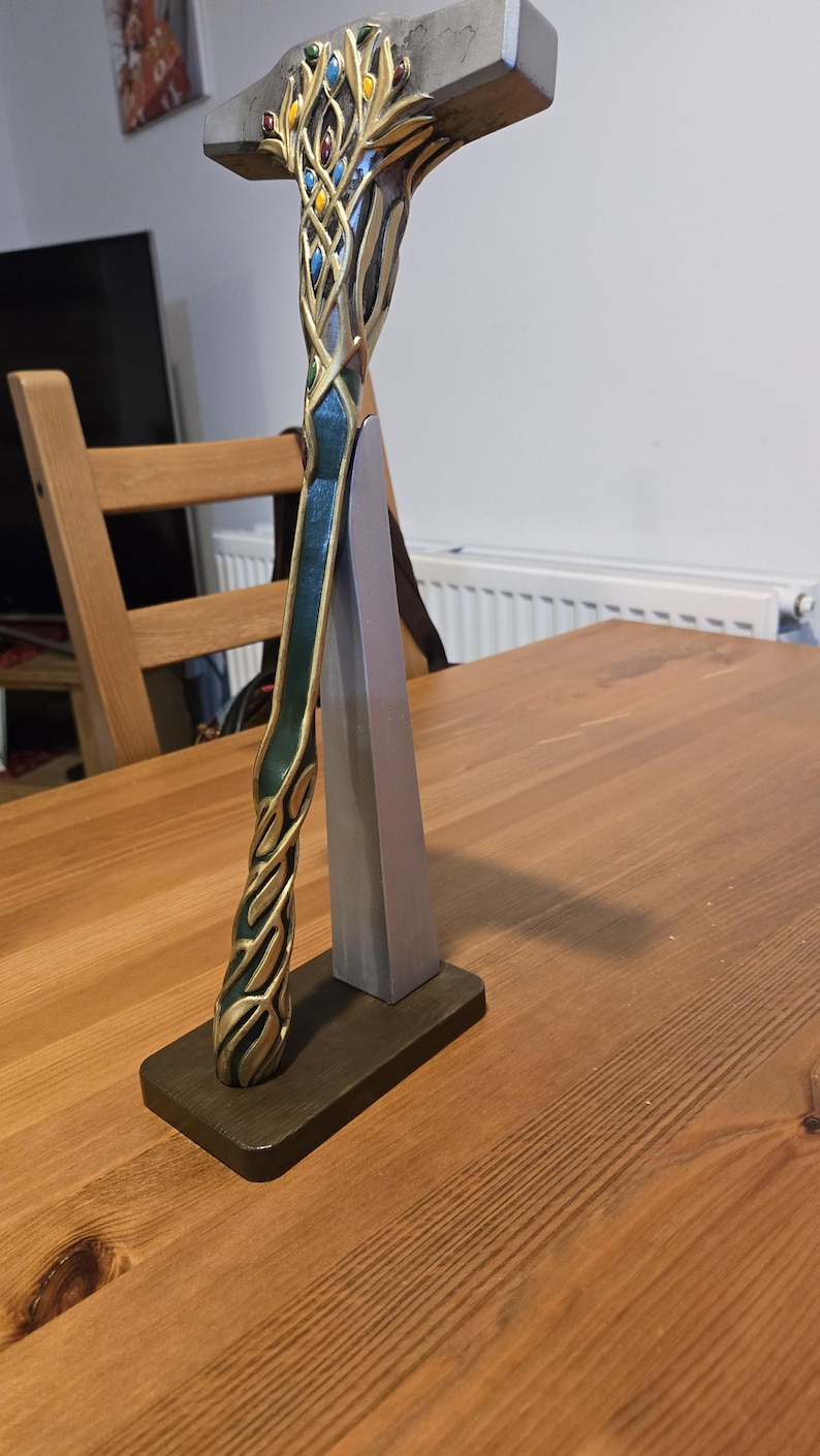 May include: A decorative hammer with a gold and green handle, featuring intricate details and colorful gems. The hammer is displayed on a wooden stand.