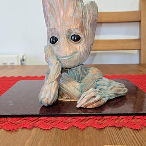 May include: A Groot-style figurine, a detailed sculpture with a textured, wood-like appearance. The figure is in a thoughtful pose, resting its chin on its hand. The colour palette includes shades of brown and teal, with black eyes.