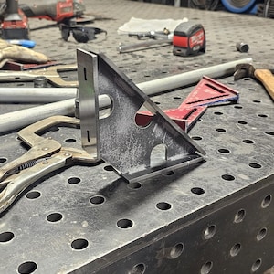 May include: A metal welding table with a variety of tools and equipment, including a metal square, a clamp, and a measuring tape. The table has a grid pattern of holes.