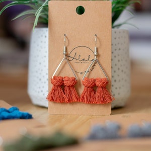 May include: A pair of orange tassel earrings with silver triangle frames. The earrings are displayed on a brown card with the text "Made with Love" written on it.