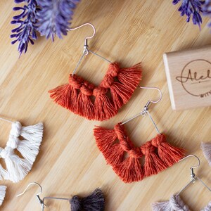 May include: A set of four pairs of handmade macrame earrings with a square shape and fringe. The earrings are in three colors: white, beige, and orange. The earrings are displayed on a wooden surface with a small wooden box that says "Alela with love".