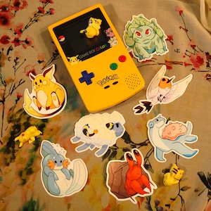 May include: A yellow Game Boy Color with a Pokemon game cartridge, surrounded by various Pokemon character stickers. The stickers include a yellow Pikachu, a green Bulbasaur, a blue Squirtle, a white and blue  Eevee, a red and brown  Krabby, a blue and white  Lapras, a yellow Pichu, and a white and blue  Flaaffy.