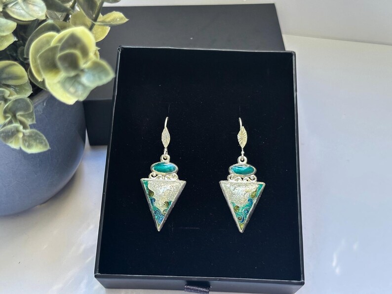 May include: A pair of silver-toned earrings with a triangular pendant. The earrings feature a teal-colored oval stone at the top and a detailed enamel design in teal, white, and silver. The earrings are displayed in a black box.