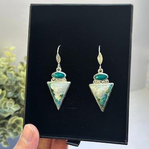 May include: A pair of silver-toned earrings with a triangular pendant design. Each earring features a green oval stone at the top and a triangle with a silver and enamel design. The earrings are displayed in a black box.