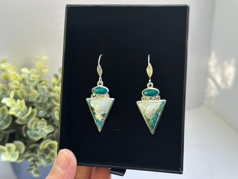 May include: A pair of silver-toned earrings with a triangular pendant design. Each earring features a green oval stone at the top and a triangle with a silver and enamel design. The earrings are displayed in a black box.