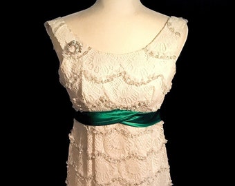 May include: A white lace dress with a green satin sash. The dress has a scalloped neckline and a fitted silhouette.