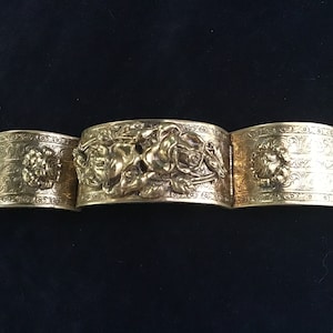 May include: A gold-toned metal bracelet with an ornate design featuring floral motifs and a central figure. The bracelet is made of three sections that are connected by hinges.