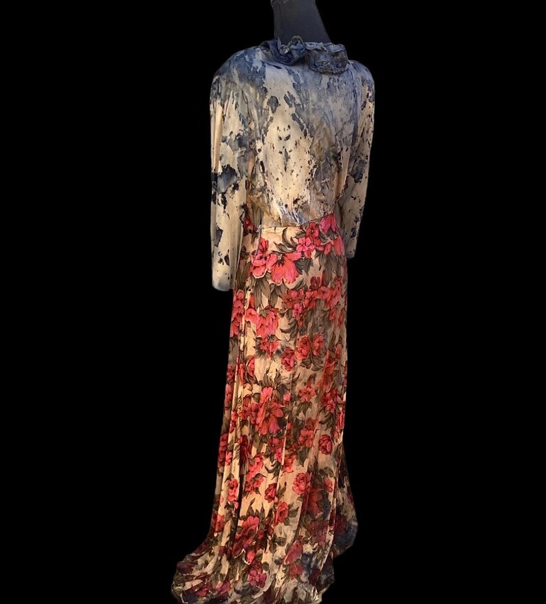 May include: A long, flowing dress with a floral print skirt and a patterned top. The top features a ruffled collar and long sleeves. The skirt is adorned with large, vibrant pink flowers against a cream background. The dress is displayed on a mannequin.